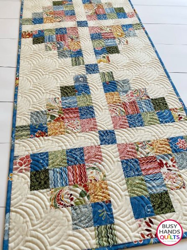 May include: A white quilted table runner with a patchwork design featuring blue, green, red, and yellow squares. The quilting pattern is a series of curved lines. The table runner has the text "Busy Hands Quilts" in the bottom right corner.