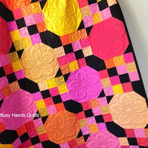 Könnte beinhalten: Ein farbenfroher Quilt mit einem geometrischen Muster aus Quadraten und Achtecken. Der Quilt ist aus verschiedenen Stoffen in den Farben Rosa, Orange, Gelb und Schwarz gefertigt. Der Quilt ist mit einem floralen Muster gesteppt. Der Text "Busy Hands Quilts" ist in der linken unteren Ecke des Quilts sichtbar.