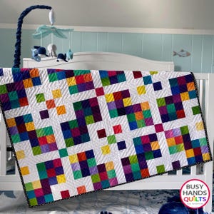 May include: A colorful patchwork quilt with a white background and a black and white polka dot border. The quilt is made up of squares of different colors, including red, orange, yellow, green, blue, purple, and pink. The quilt is hanging on a white crib with a blue and white wall behind it. The quilt is made by Busy Hands Quilts.