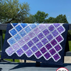 Puede incluir: Una colcha de color morado y azul con un patrón geométrico de cuadrados y círculos. La colcha está colgada en una barandilla de madera con un césped verde y árboles en el fondo. La colcha está hecha por Busy Hands Quilts, que está impreso en una etiqueta adjunta a la colcha.