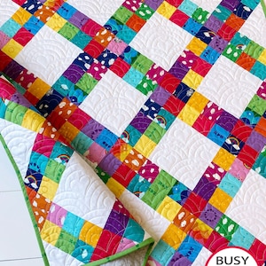 Puede incluir: Una colcha de retazos colorida con fondo blanco y borde verde. La colcha está hecha de cuadrados de diferentes colores y patrones, incluyendo arcoíris, nubes y corazones. La colcha está doblada por la mitad y la esquina está levantada para mostrar la parte posterior de la colcha, que es una tela blanca sólida. La colcha está etiquetada como "Busy Hands Quilts" en la esquina inferior derecha.