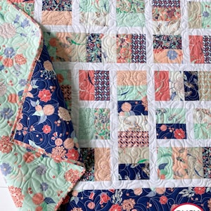 May include: A patchwork quilt featuring rectangular blocks in shades of coral, mint green, and navy blue with floral and bow-and-arrow patterns. The quilt has white sashing and a navy blue floral border. A logo reads BUSY HANDS QUILTS buy handmade.