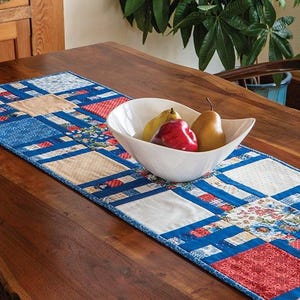 Könnte beinhalten: Ein blauer, roter und weißer Patchwork-Tischläufer mit einer weißen Schüssel mit Obst auf einem Holztisch. Der Tischläufer hat ein kariertes Muster und der Text "Busy Hands Quilts" befindet sich in der rechten unteren Ecke.