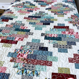 May include: A white quilt with a patchwork design featuring various shades of blue, green, red, and pink. The quilt is made with a variety of fabrics, including floral prints, geometric patterns, and solid colors. The quilt is quilted with a swirling pattern. The quilt is labeled "Busy Hands Quilts" in the bottom right corner.