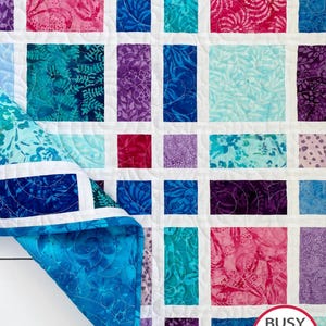 May include: A handmade quilt with a grid pattern of colorful fabric squares in shades of teal, blue, purple, and pink, separated by white borders. The quilt's underside is a vibrant teal with a swirling pattern. The quilt is labeled "Busy Hands Quilts".