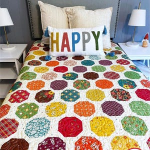 Może przedstawiać: Białe łóżko z szarym zagłówkiem i kolorową patchworkową kołdrą. Kołdra zawiera różne wzory kwiatowe i geometryczne w odcieniach czerwieni, pomarańczy, żółci, zieleni i błękitu. Biała poduszka z napisem "HAPPY" w kolorowych literach leży na kołdrze.