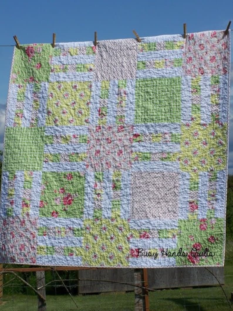 May include: A patchwork quilt with a green, white, and pink floral pattern. The quilt is hanging on a clothesline with wooden clothespins. The quilt is labeled "Busy Hands Quilts".
