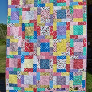 May include: A colorful patchwork quilt with squares of various floral and geometric patterns in shades of pink, yellow, blue, green, and purple. The quilt is hanging on a clothesline and has the text "Busy Hands Quilts" in the bottom right corner.