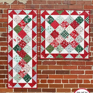 May include: Two Christmas-themed quilts with red, green, and white patchwork designs. The quilts feature diamond patterns and floral prints, with a red border. One is rectangular, the other square. The quilts are displayed against a brick wall.