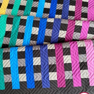 Pode incluir: Um cobertor colorido com tecido xadrez preto e branco e faixas de diferentes cores, incluindo amarelo, verde, azul e roxo. O cobertor é acolchoado com um padrão de chevron. O cobertor é etiquetado como "Busy Hands Quilts" no canto inferior direito.