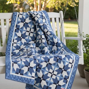 Puede incluir: Una colcha con un patrón de estrellas geométricas en tonos azul marino, azul claro y blanco. La tela tiene un borde floral y un logotipo circular en la esquina que dice BUSY HANDS QUILTS. La colcha está sobre un banco de madera blanco.