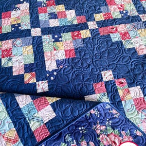 May include: A blue and white quilt with a floral pattern. The quilt is made up of small squares and is quilted with a swirl pattern. The quilt is folded in half and the bottom edge is visible. The quilt is labeled "Busy Hands Quilts" in the bottom right corner.