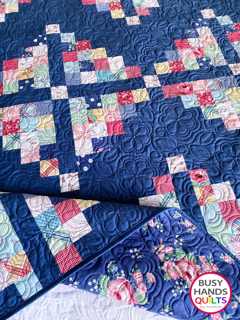 May include: A blue and white quilt with a floral pattern. The quilt is made up of small squares and is quilted with a swirl pattern. The quilt is folded in half and the bottom edge is visible. The quilt is labeled "Busy Hands Quilts" in the bottom right corner.