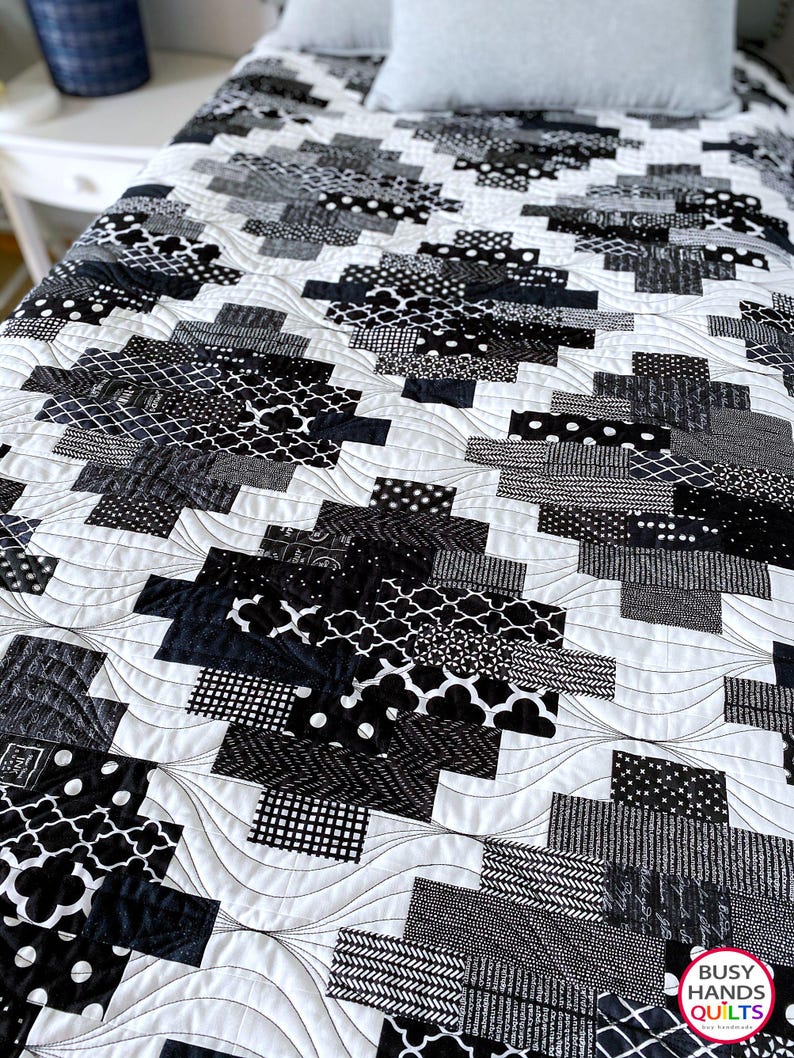 May include: Black and white quilt with a geometric pattern. The quilt is made of various fabrics with different black and white patterns. The quilt is quilted with a wavy pattern. The quilt is on a bed with a white pillow.