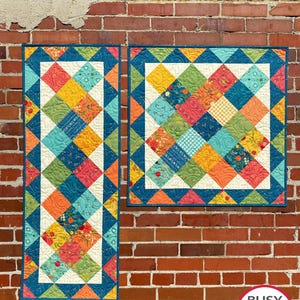 Könnte beinhalten: Zwei handgefertigte Quilts mit einem geometrischen Rautenmuster. Die Quilts weisen eine Vielzahl von Stoffen in Blau-, Orange-, Gelb- und Grüntönen auf. Die Quilts werden vor einer Backsteinmauer ausgestellt. Die Quilts sind mit "Busy Hands Quilts" beschriftet.
