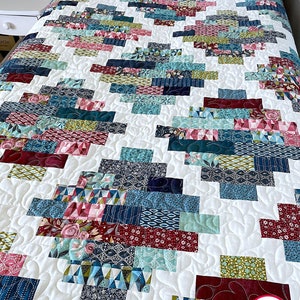 May include: A white quilt with a patchwork design featuring various shades of blue, green, red, and pink. The quilt is made with a variety of fabrics, including floral prints, geometric patterns, and solid colors. The quilt is quilted with a swirling pattern. The quilt is labeled "Busy Hands Quilts" in the bottom right corner.