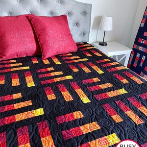 May include: A black quilt with a swirling pattern and red, orange, and yellow squares. The quilt is on a bed with red pillows and a gray headboard. The text "Busy Hands Quilts" is in a circle on the bottom right corner of the quilt.