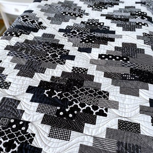 May include: Black and white quilt with a geometric pattern. The quilt is made of various fabrics with different textures and patterns. The quilt is quilted with a wavy design. The quilt is on a bed with a white pillow.