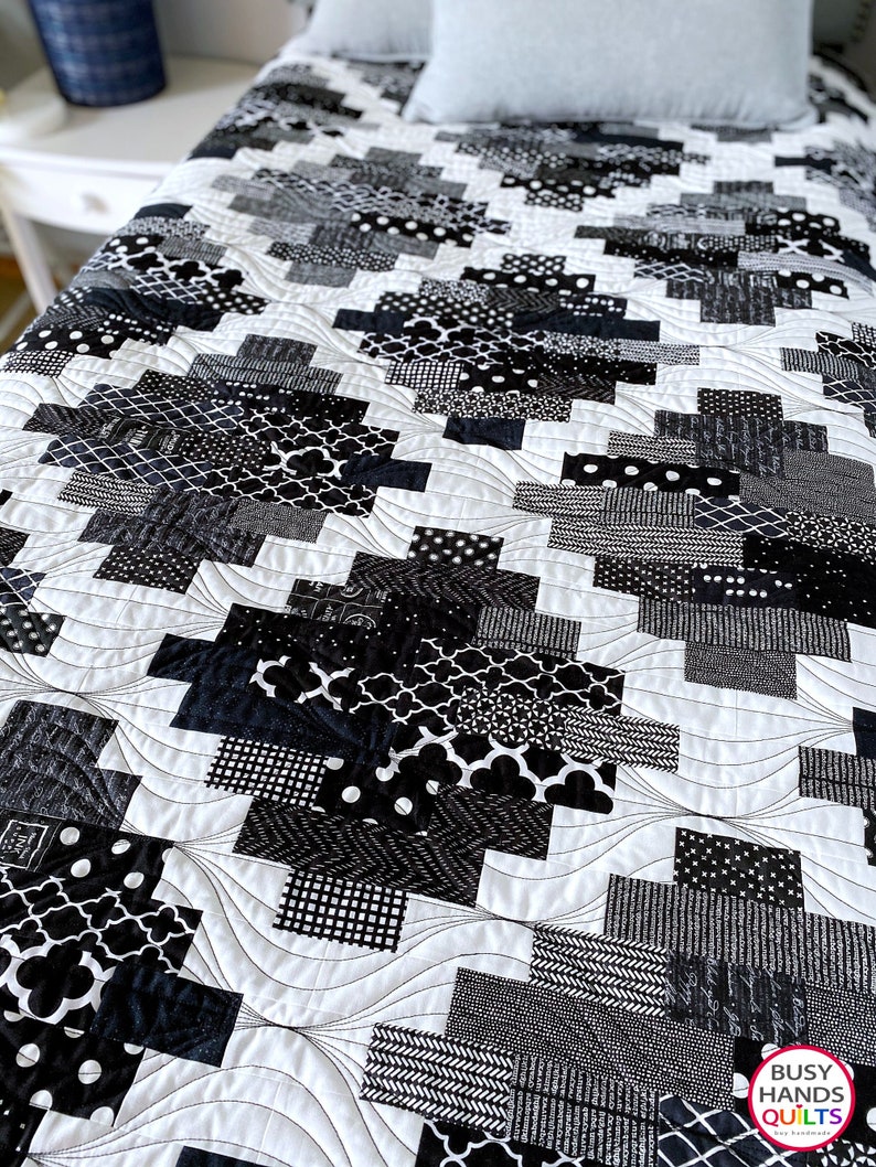 May include: Black and white quilt with a geometric pattern. The quilt is made of various fabrics with different textures and patterns. The quilt is quilted with a wavy design. The quilt is on a bed with a white pillow.