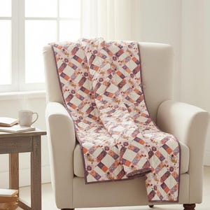 Op de afbeelding: Een gewatteerde sprei met een diagonaal patchworkpatroon van witte, oranje en paarse bloemrijke stoffen vierkanten. De quilt heeft een effen paarse stoffen rand en is gedrapeerd over een crèmekleurige fauteuil. Een logo toont Busy Hands Quilts.