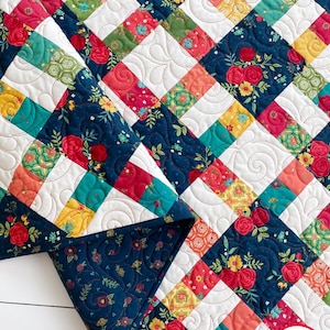 May include: A patchwork quilt featuring a grid of squares in navy blue, white, teal, yellow, and red. The fabric displays floral patterns with roses and small blossoms. The white squares feature a swirling quilted texture. Text reads BUSY HANDS QUILTS.
