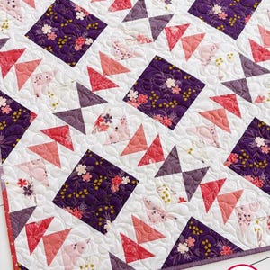 May include: A purple and pink quilt with a geometric pattern of triangles and squares. The quilt is made with a floral fabric and has a white background. The quilt is handmade and has the text "Busy Hands Quilts" in the bottom right corner.