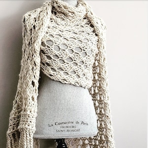 May include: A cream-coloured crocheted shawl with fringe on a mannequin. The shawl has a lacy pattern and is draped around the mannequin's shoulders. The mannequin is wearing a grey dress and has a black stand. The label on the mannequin reads "La Couturiere de Paris, Faubourg Saint-Honore."