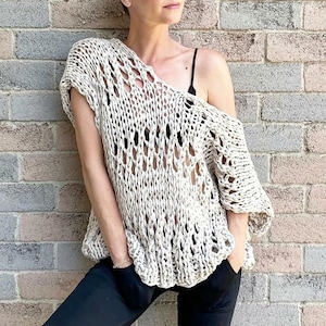 May include: A loose-fitting, off-white knitted top with a large open-weave pattern. The top has short sleeves and a relaxed fit.
