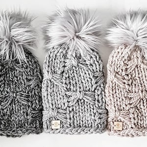 May include: Three knitted winter hats with faux fur pom poms. The hats are in gray, light gray, and beige. Each hat has a small leather tag with the word "Cozy" on it.