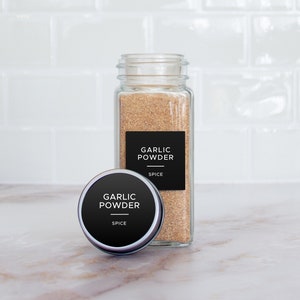 Op de afbeelding: Een glazen pot gevuld met knoflookpoeder, met een zwart etiket met de tekst "Garlic Powder" en "Spice". Naast de pot staat een kleinere, ronde metalen container met een vergelijkbaar etiket.