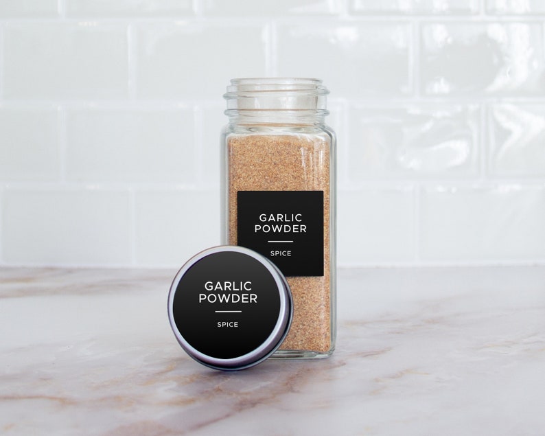 Op de afbeelding: Een glazen pot gevuld met knoflookpoeder, met een zwart etiket met de tekst "Garlic Powder" en "Spice". Naast de pot staat een kleinere, ronde metalen container met een vergelijkbaar etiket.