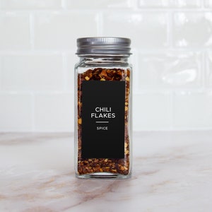 Op de afbeelding: Een glazen pot met een zilveren deksel gevuld met rode chilivlokken. De pot heeft een zwart etiket met de tekst "Chili Flakes" en "Spice".