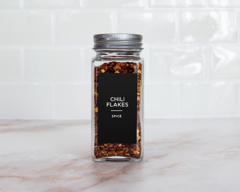 Op de afbeelding: Een glazen pot met een zilveren deksel gevuld met rode chilivlokken. De pot heeft een zwart etiket met de tekst "Chili Flakes" en "Spice".