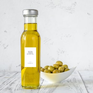 May include: A clear glass bottle of extra virgin olive oil with a white label that reads "Extra Virgin Olive Oil". The bottle is sitting on a white wooden surface with a bowl of green olives in front of it.