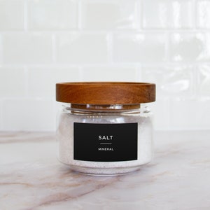 Op de afbeelding: Een glazen pot met een houten deksel gevuld met wit zout. De pot heeft een zwart etiket met de tekst "SALT MINERAL".