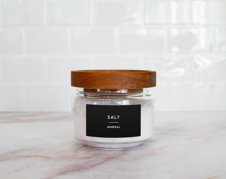 Op de afbeelding: Een glazen pot met een houten deksel gevuld met wit zout. De pot heeft een zwart etiket met de tekst "SALT MINERAL".