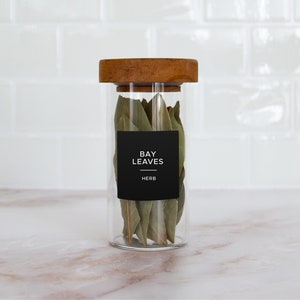 Op de afbeelding: Een glazen pot met een houten deksel gevuld met gedroogde laurierbladeren. De pot heeft een zwart etiket met de tekst "BAY LEAVES HERB".