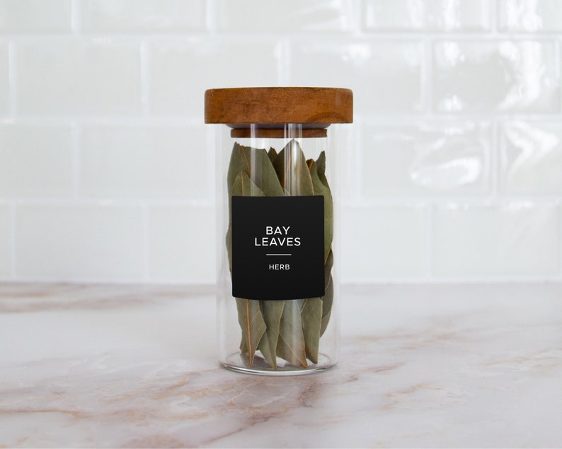 Op de afbeelding: Een glazen pot met een houten deksel gevuld met gedroogde laurierbladeren. De pot heeft een zwart etiket met de tekst "BAY LEAVES HERB".