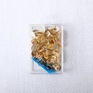 May include: A clear plastic box filled with numerous gold-colored push pins. The pins have a flat, round head and a sharp point. A small blue plastic piece is visible at the bottom of the box. The background is a white, textured surface.