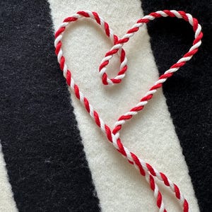 May include: A heart shape made of red and white twisted cord against a black and white striped background. The cord is tightly woven, creating a textured appearance. The heart is centered, with the cord extending downwards.