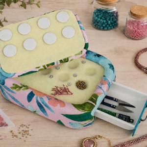 May include: A floral patterned fabric bead storage box with a hinged lid featuring eight white circular pads. The interior contains a foam insert with circular indentations for beads and a pull-out white plastic drawer containing small metal scissors.