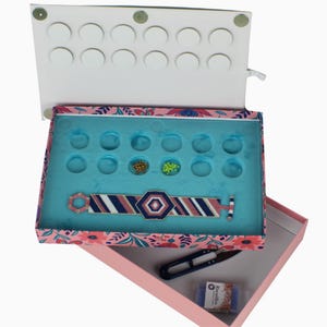 May include: A rectangular box with a floral pattern containing a blue tray with circular indentations, a beaded bracelet with a geometric design in navy, pink, and white, a pair of dark blue thread snips, and a small container labelled Rocailles.
