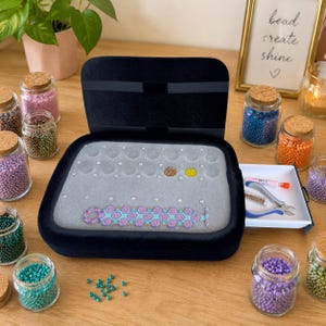 May include: A black jewelry box with an open lid, revealing a gray interior with a beaded bracelet and various beads. Small glass jars filled with colorful beads surround the box, along with tools.