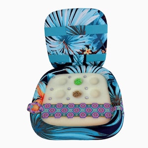 May include: A portable bead loom case featuring a blue and teal tropical leaf print fabric. The interior contains a white foam insert with six circular indentations labeled A through I, holding small piles of green and gold beads and a woven band.