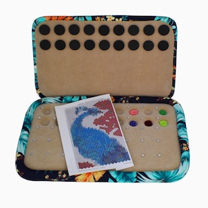 May include: A rectangular open storage case with a dark floral fabric exterior featuring teal, orange, and yellow flowers. The interior has a beige foam insert with circular cutouts for beads, small metal pins, and a printed peacock pattern chart.