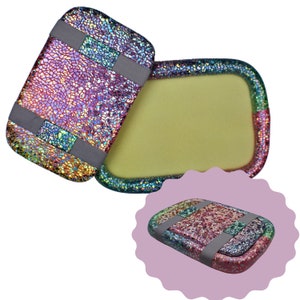 May include: Two rectangular cases with a vibrant, iridescent, mosaic-like finish in shades of pink, blue, and gold. One case is open, revealing a smooth, pale yellow interior, while the other is closed with two grey elastic bands across it.