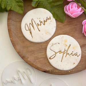 May include: Two white round cake toppers with gold lettering. The toppers say "Sophia" and "Marina". Two white round silicone molds with the same names are also shown.