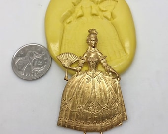 Puede incluir: Una figurita de metal dorada de una mujer con un vestido largo sosteniendo un abanico. La figurita es del tamaño aproximado de una moneda de 20 céntimos de euro.