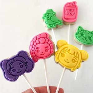 May include: A collection of colorful character-shaped cookies on sticks. The cookies feature various designs, including a bird, a soda can, a cat, and several cartoon faces in shades of pink, yellow, and purple. The cookies are on white sticks.