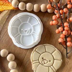 Op de afbeelding: Een witte plastic koekjesvorm en een gebakken koekje in de vorm van een leeuw. De koekjesvorm staat op een houten oppervlak met decoratieve houten kralen en oranje bessen.